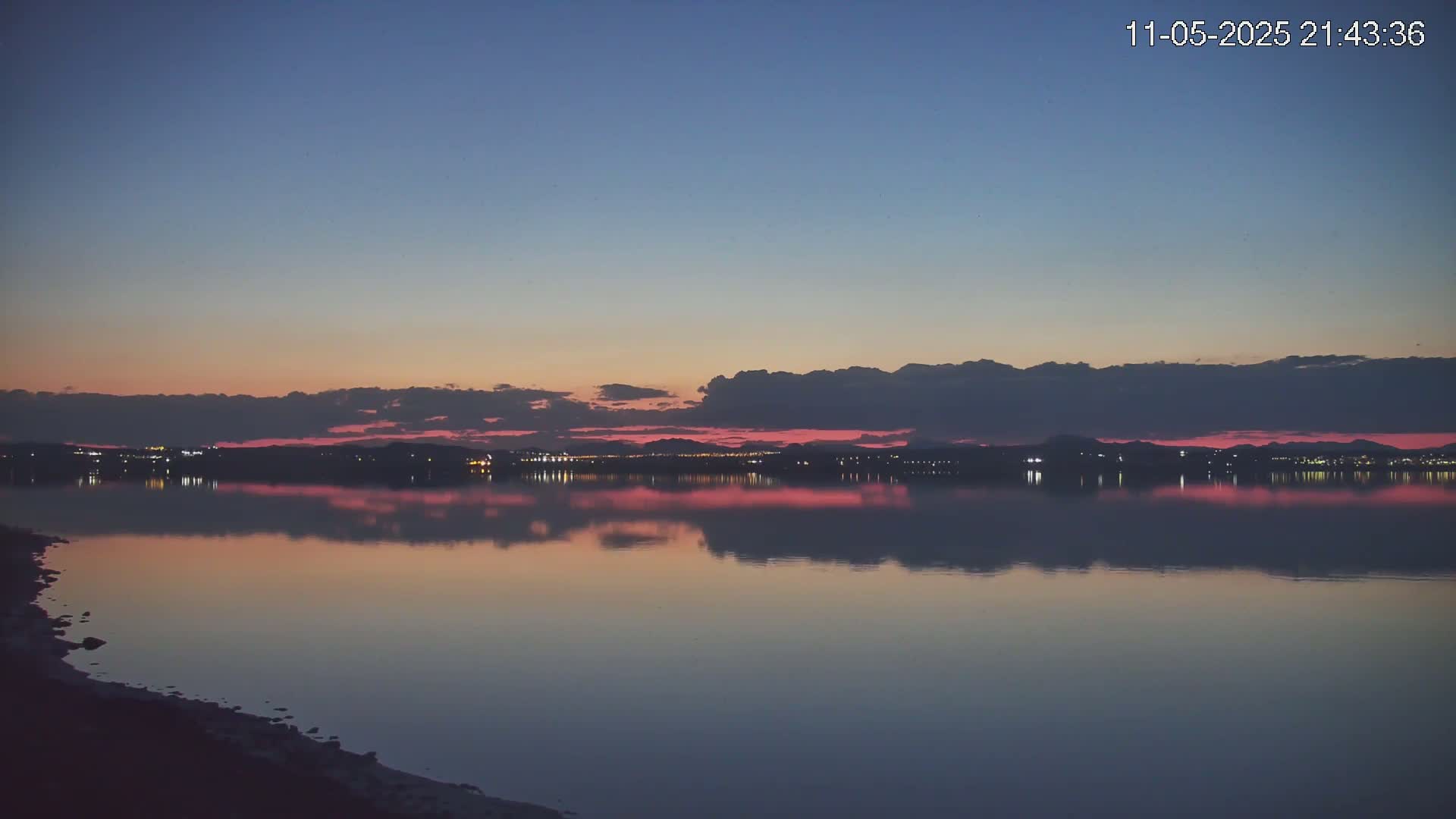 A calm body of water reflects a pink and purple sunset over a distant, dimly lit city skyline under a twilight sky.