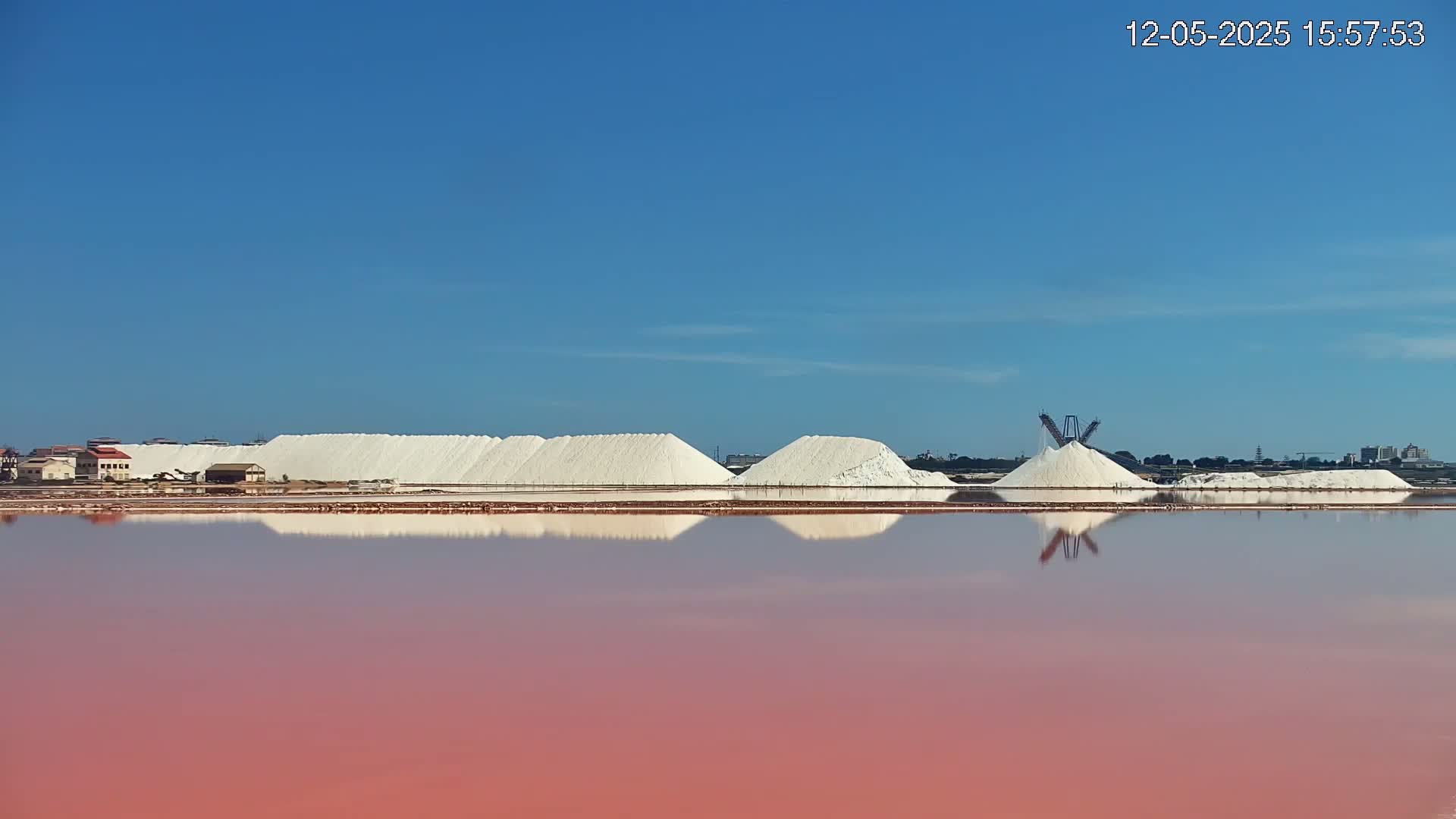 Under a clear blue sky, large piles of white salt are reflected in a pink body of water.