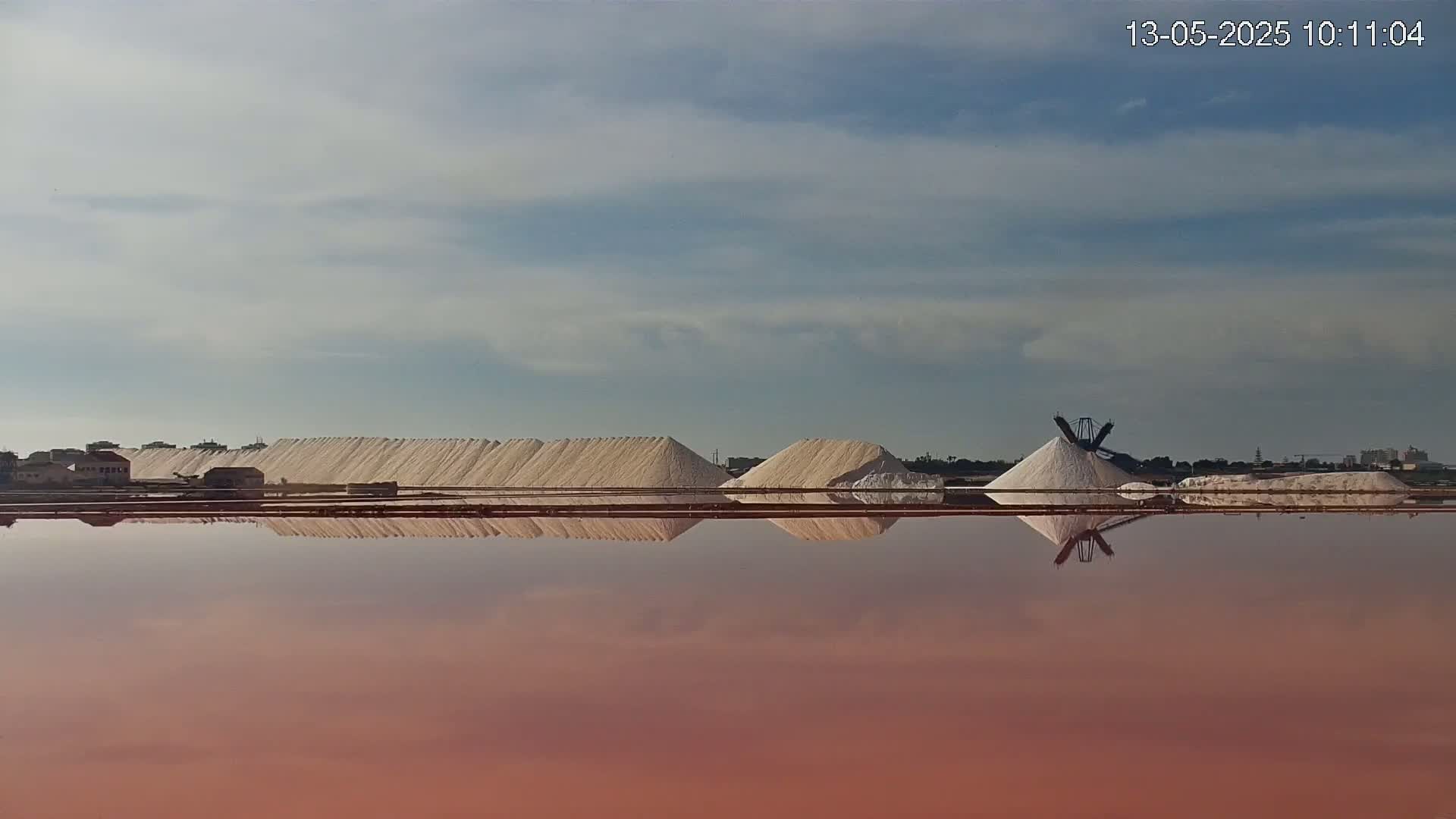 Large piles of salt are reflected in a reddish-pink body of water under a partly cloudy sky.