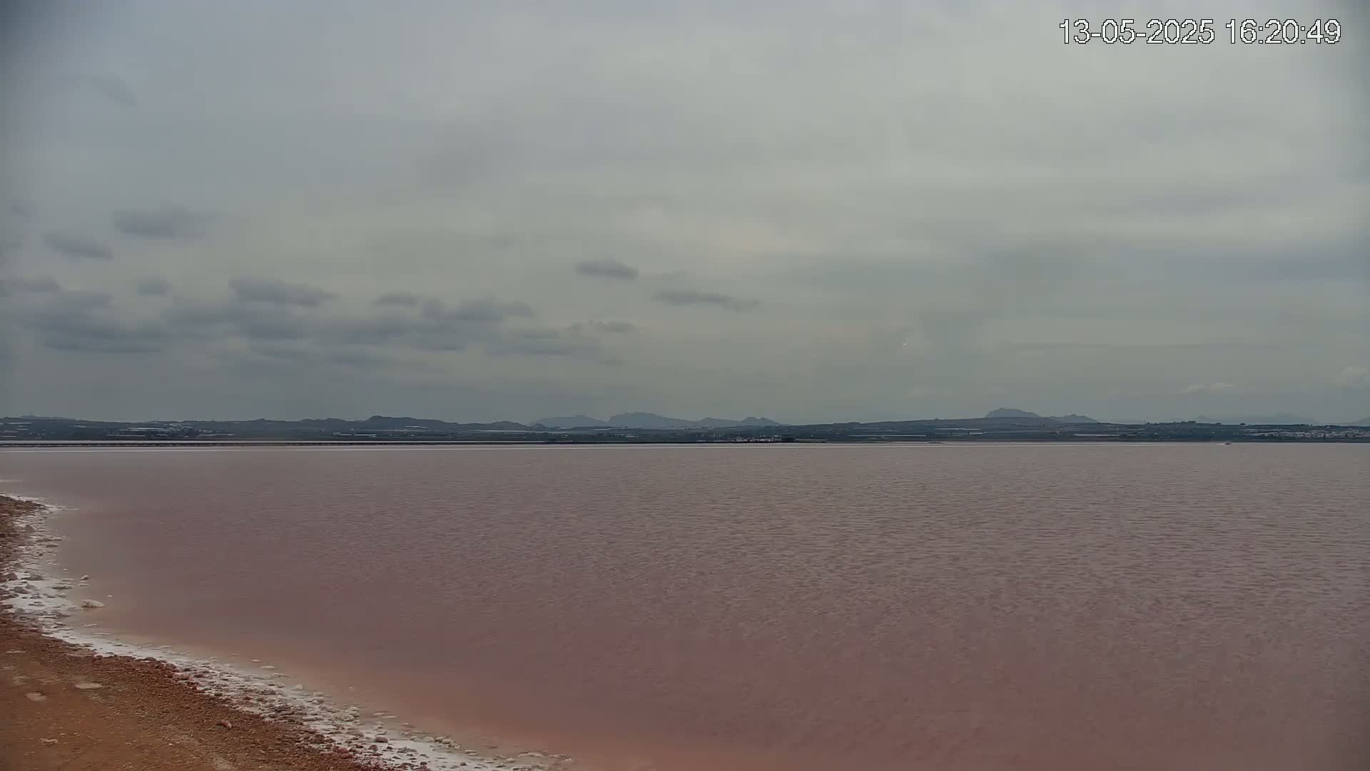 A pinkish-red body of water stretches to a distant, low-lying landmass under a cloudy sky.