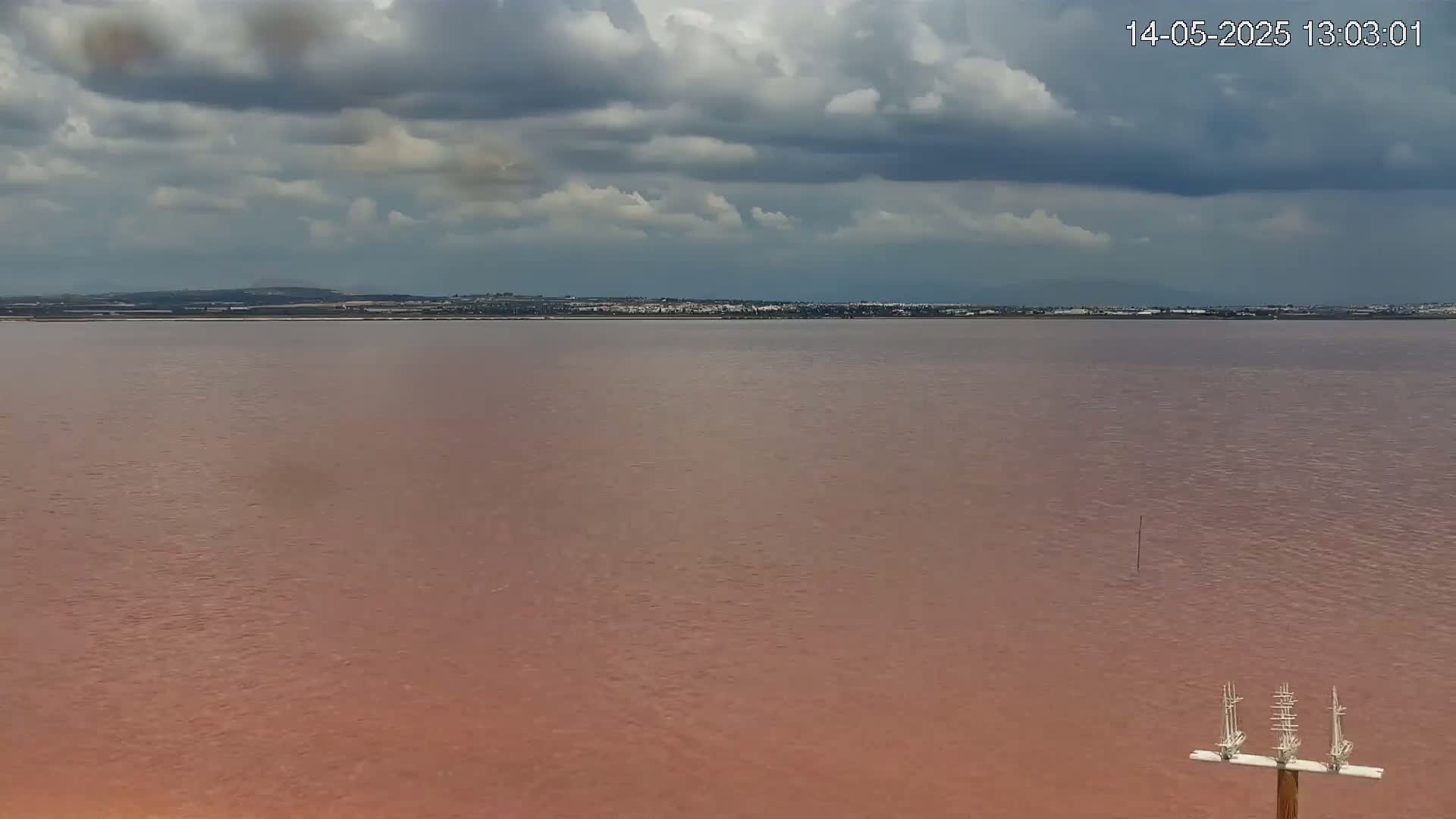 A pinkish-red body of water stretches to a distant shoreline under a mostly cloudy sky.