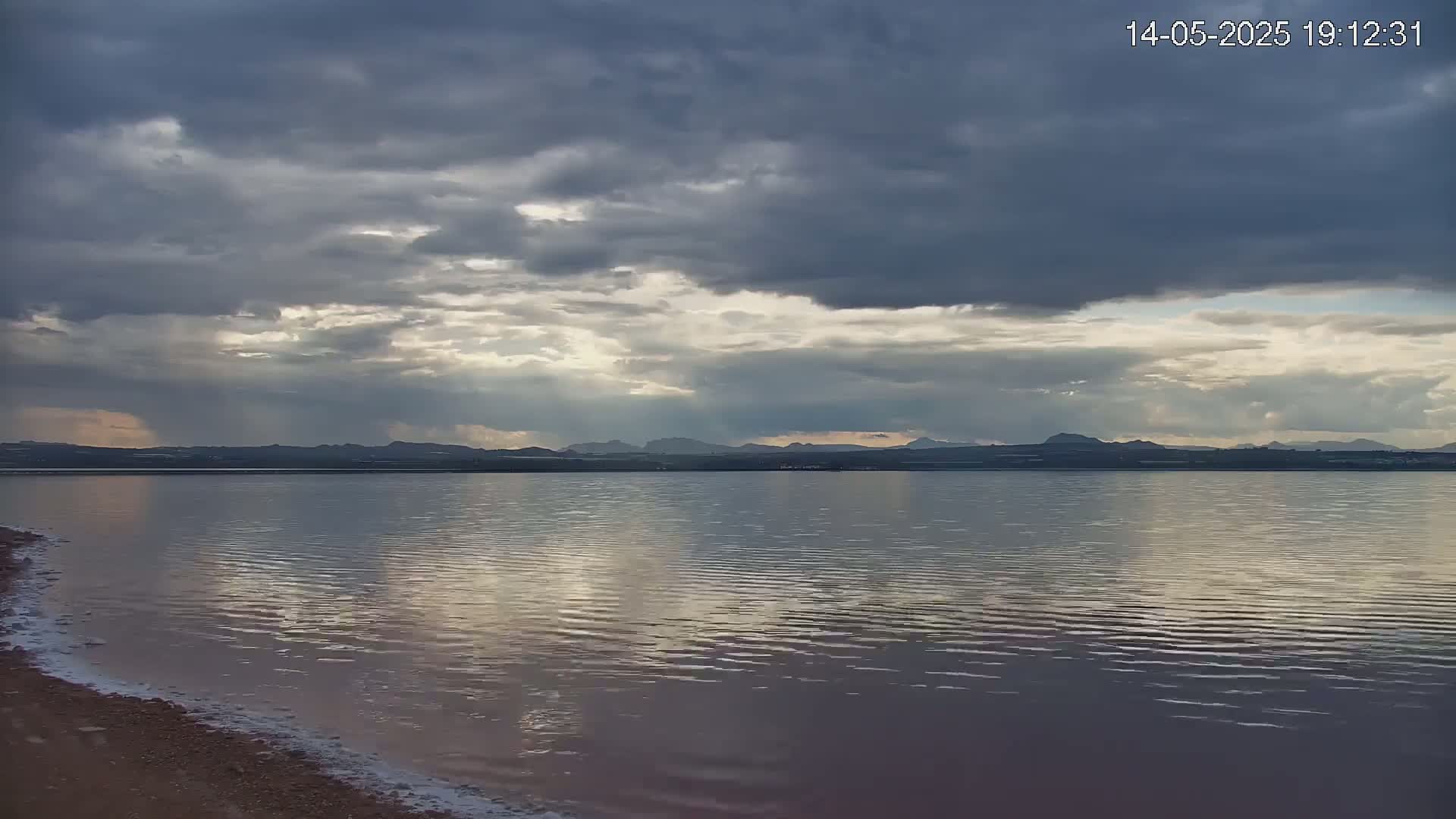 A calm body of water reflects a cloudy sky, with distant mountains visible on the horizon under a mostly overcast sky with streaks of sunlight.