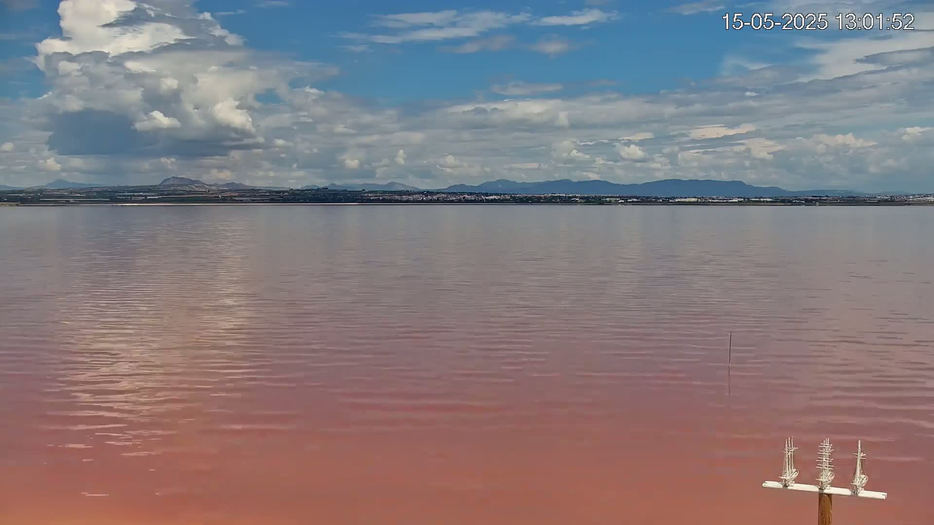 A pinkish-red lake stretches to a distant shoreline of low mountains under a partly cloudy sky.
