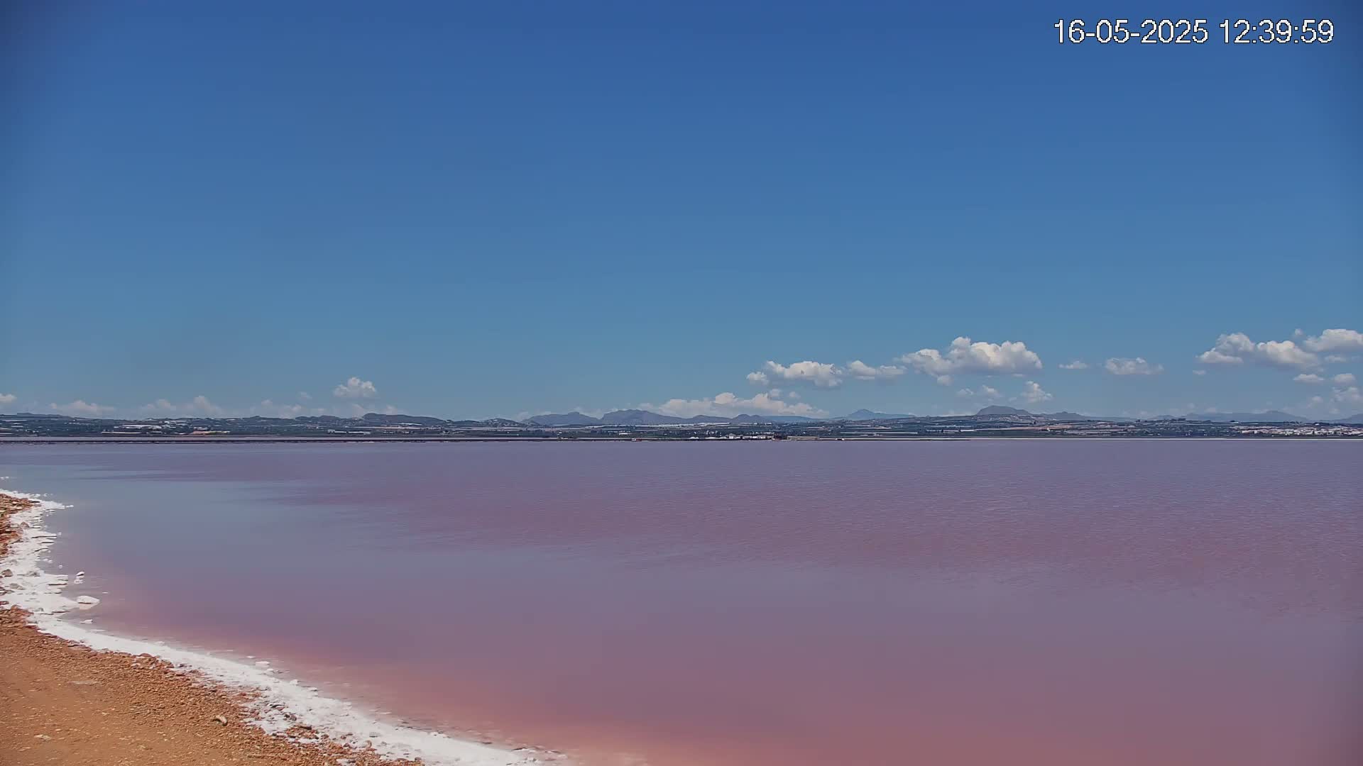 A pinkish lake stretches to a distant line of hills under a clear blue sky with a few fluffy clouds.
