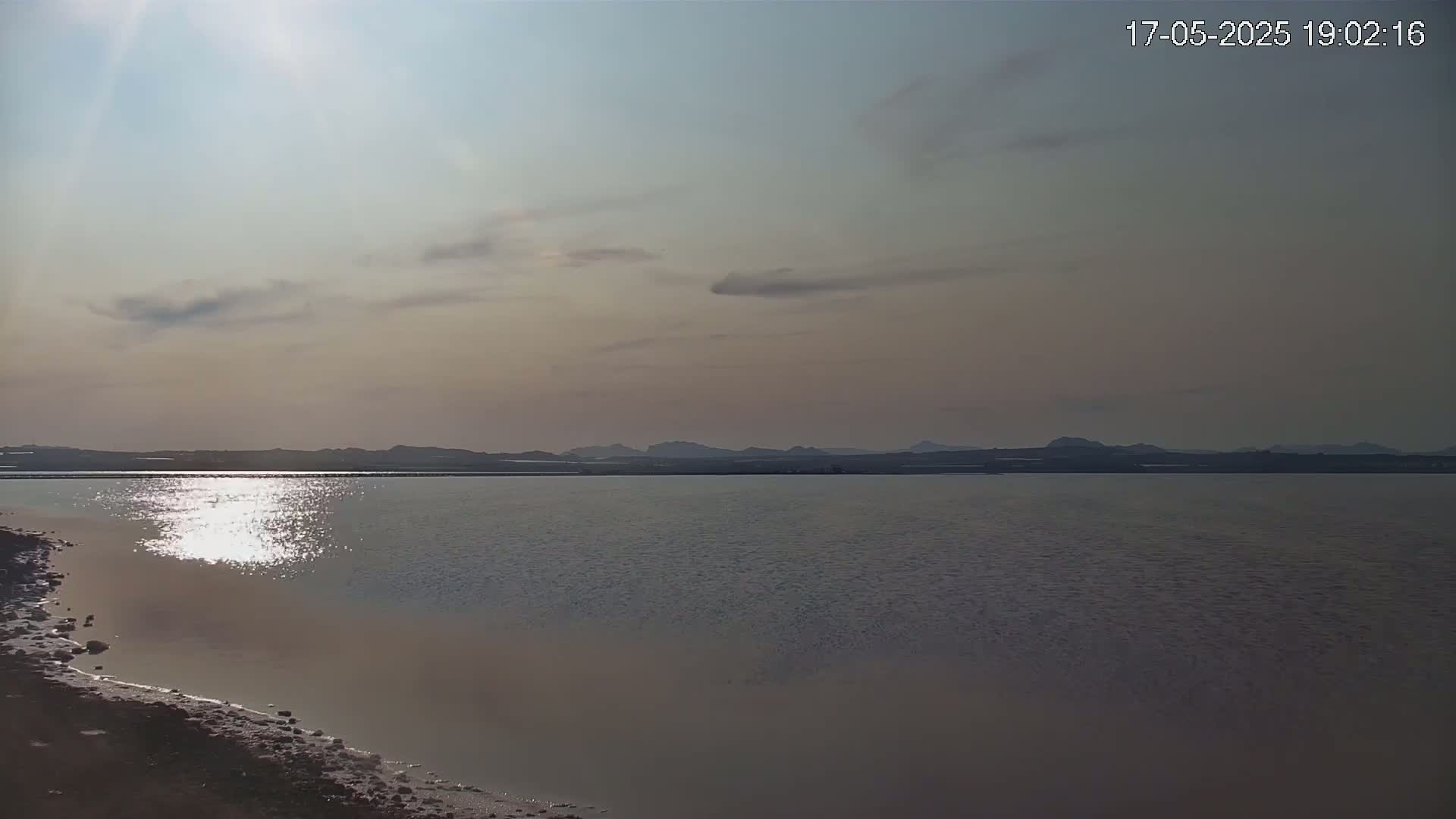 The sun shines brightly over a calm body of water with a distant shoreline under a partly cloudy sky.
