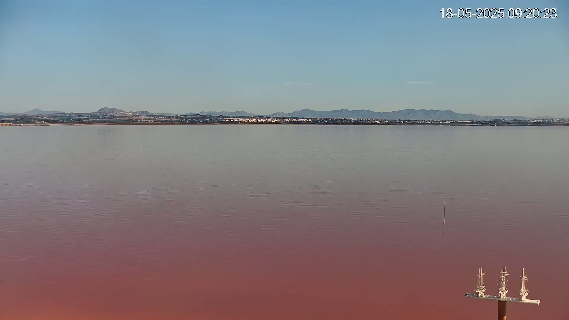 A calm, pink lake stretches to a distant shoreline of low hills under a clear blue sky.