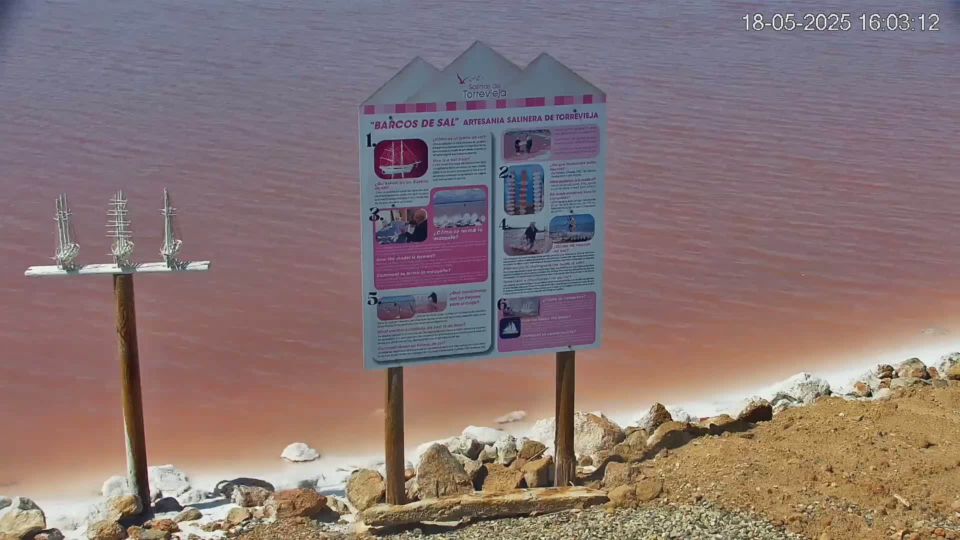 A pink-hued body of water with a white, salt-encrusted shoreline has a wooden sign with illustrations and a wooden post with three miniature sailboats.
