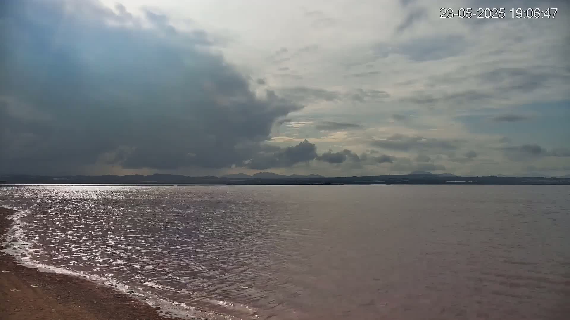 A calm body of water with a sandy shore stretches to a distant, low-lying landmass under a mostly cloudy sky, with some brighter patches visible through the clouds.