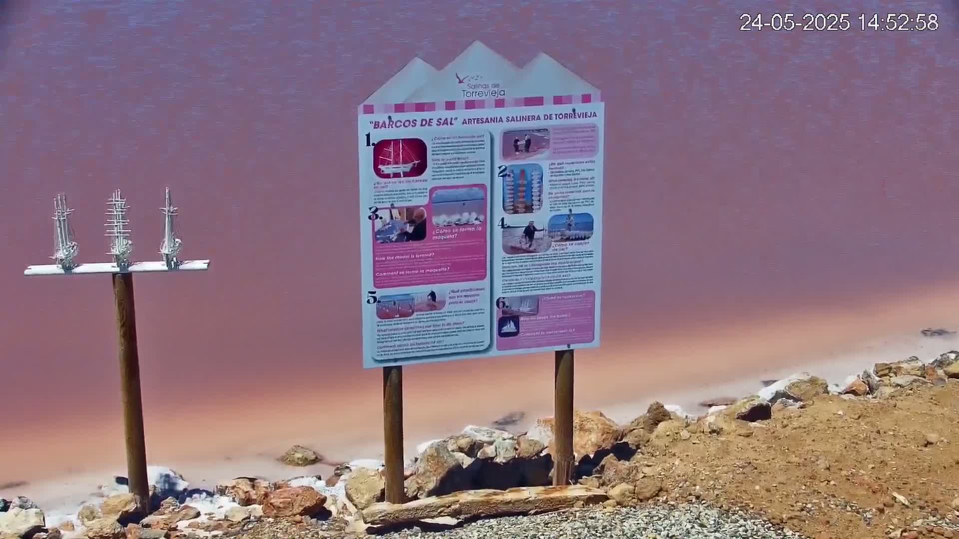 Three small model sailing ships are mounted on a post next to an informational sign by a pink lake.