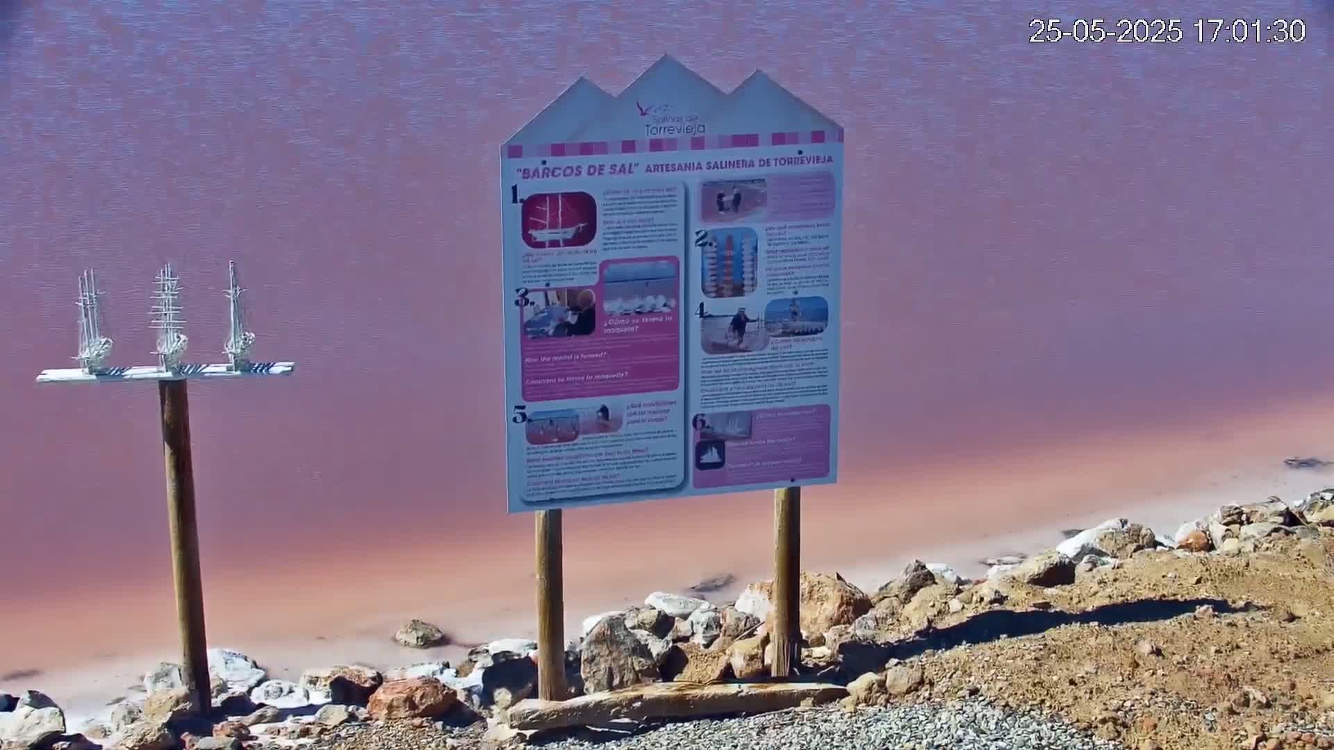 A pink salt lake is visible behind a wooden sign with illustrations and a small wooden post holding miniature white sailboats.