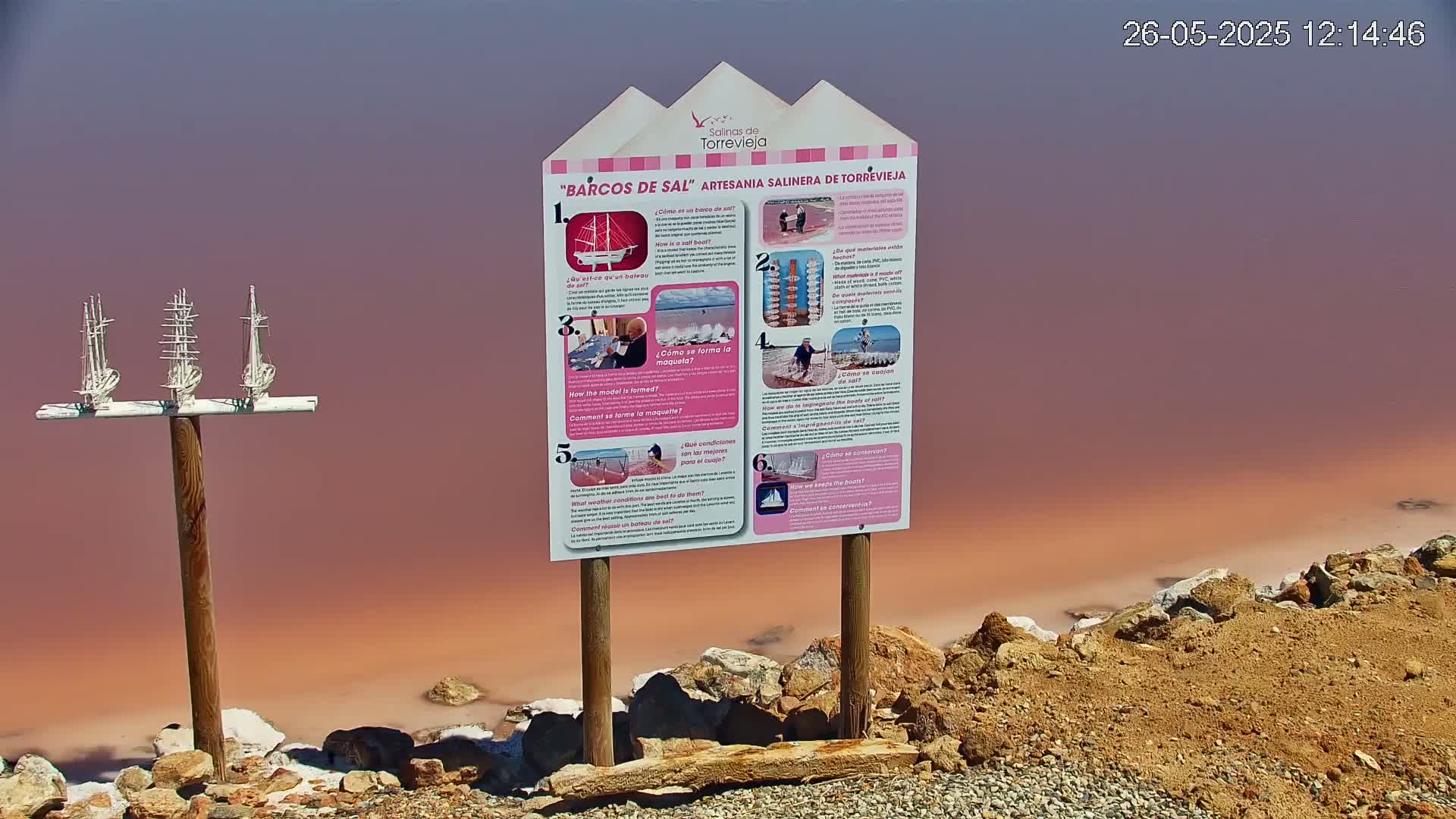 Three miniature white sailboats are mounted on a post next to an informational sign, overlooking a pink salt lake.