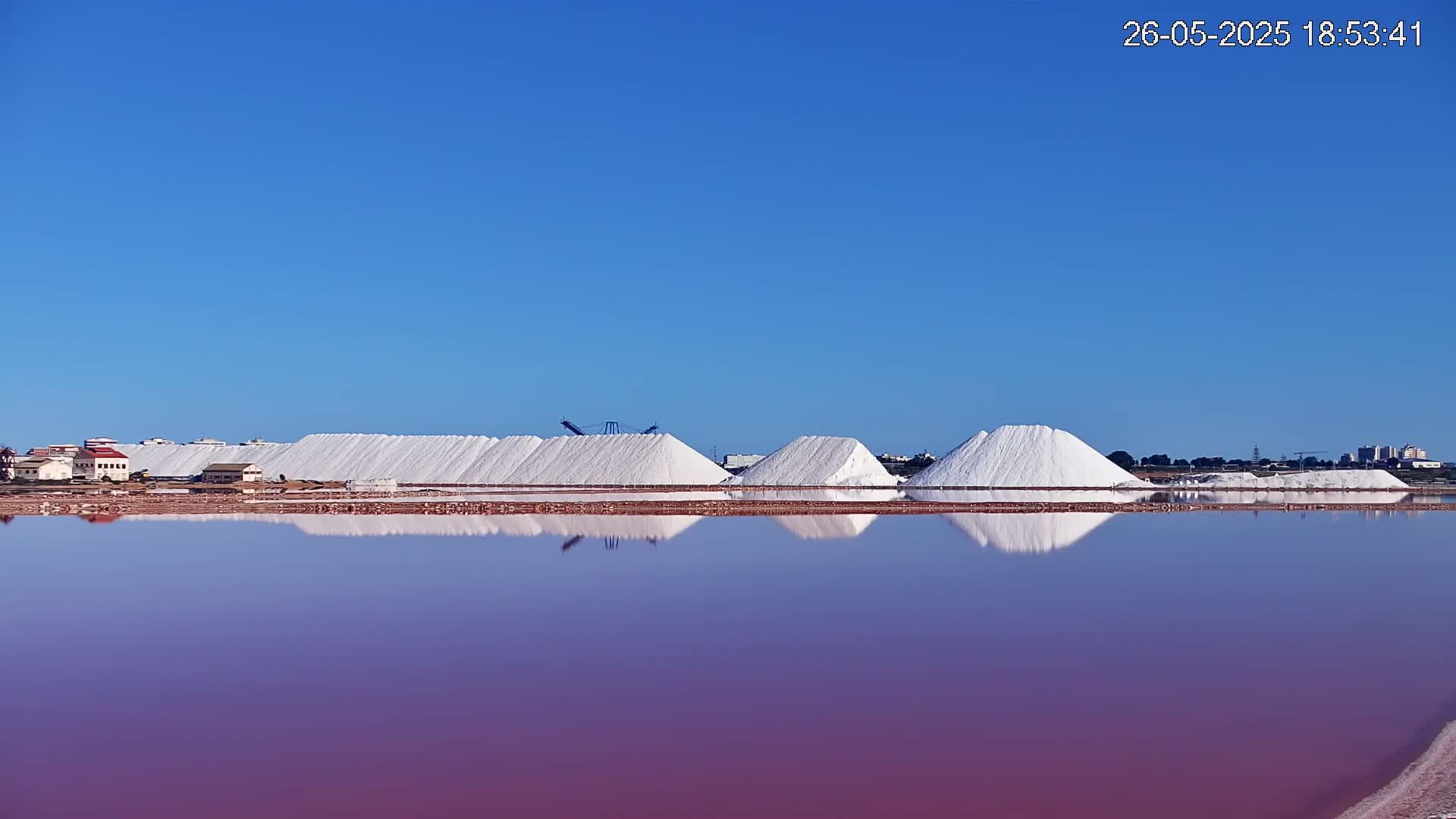 Under a clear blue sky, several large piles of white salt are reflected in a calm, purplish-pink lake.