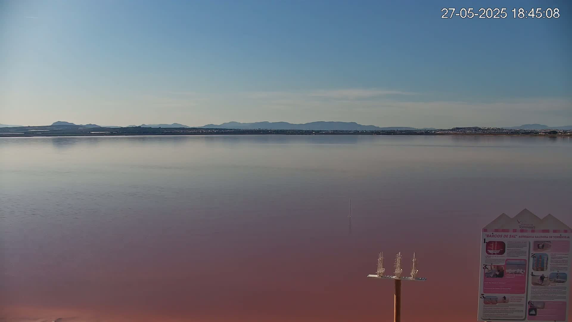 A calm, pinkish lake stretches to a distant mountain range under a clear, light blue sky.