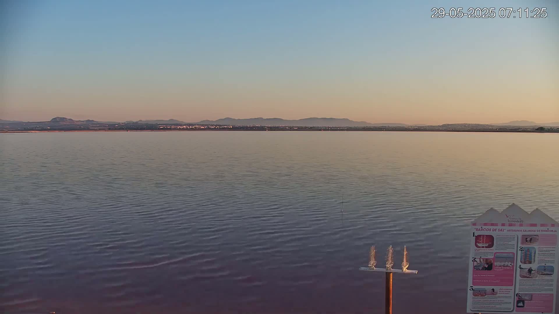 A calm body of water stretches towards a distant mountain range under a clear, pale-pink sunset sky.