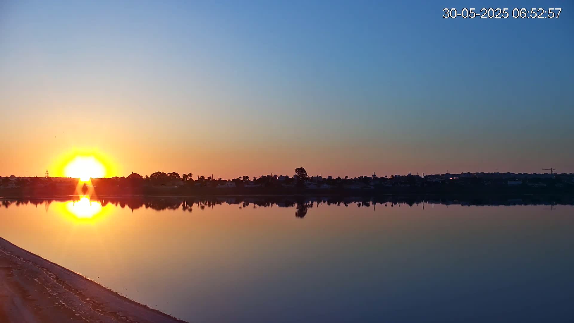 A calm lake reflects a vibrant sunrise over a distant town silhouetted against the clear, colorful sky.