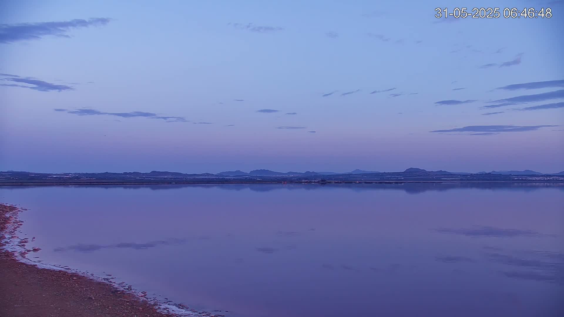A calm, purple-hued lake reflects a distant, low mountain range under a twilight sky with a few scattered clouds.