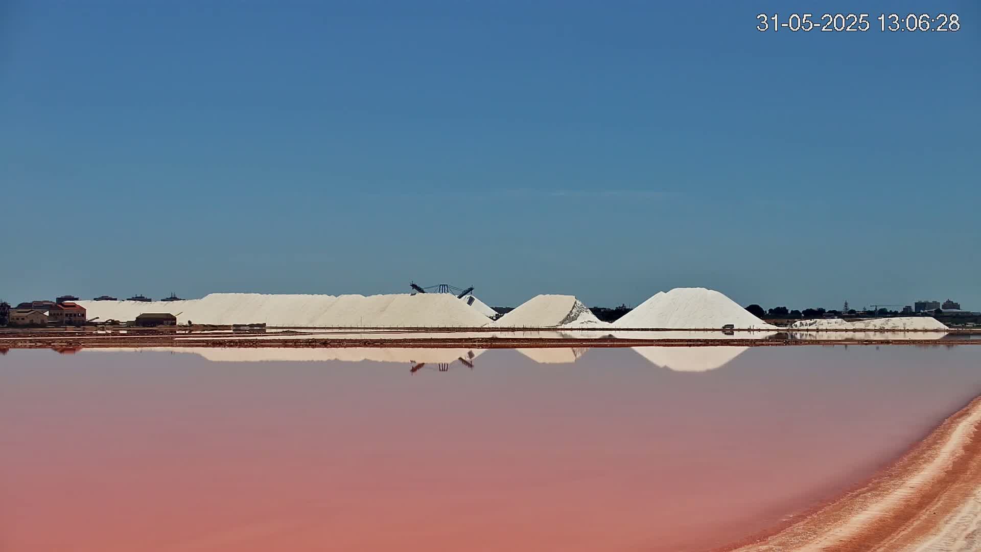 Under a clear blue sky, large piles of white salt are reflected in a pink body of water.