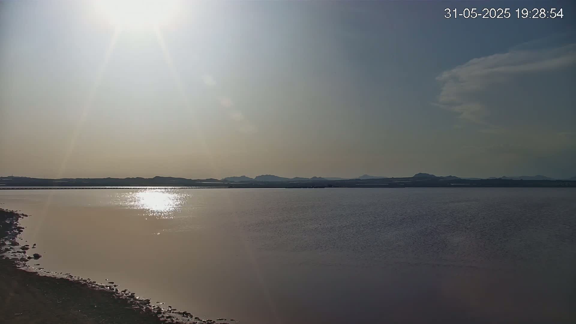 A calm body of water reflects the bright sun, with dark silhouettes of distant hills under a mostly clear sky.