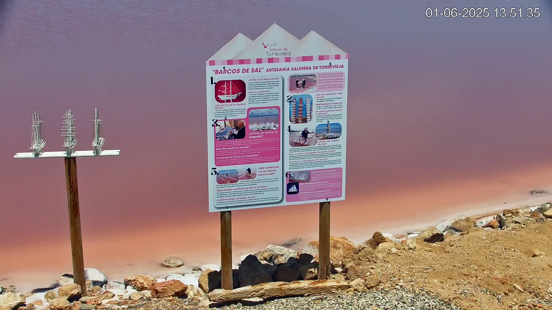 A pink salt lake shore features a wooden post with three miniature model sailing ships and an informational sign about salt boat craftsmanship.
