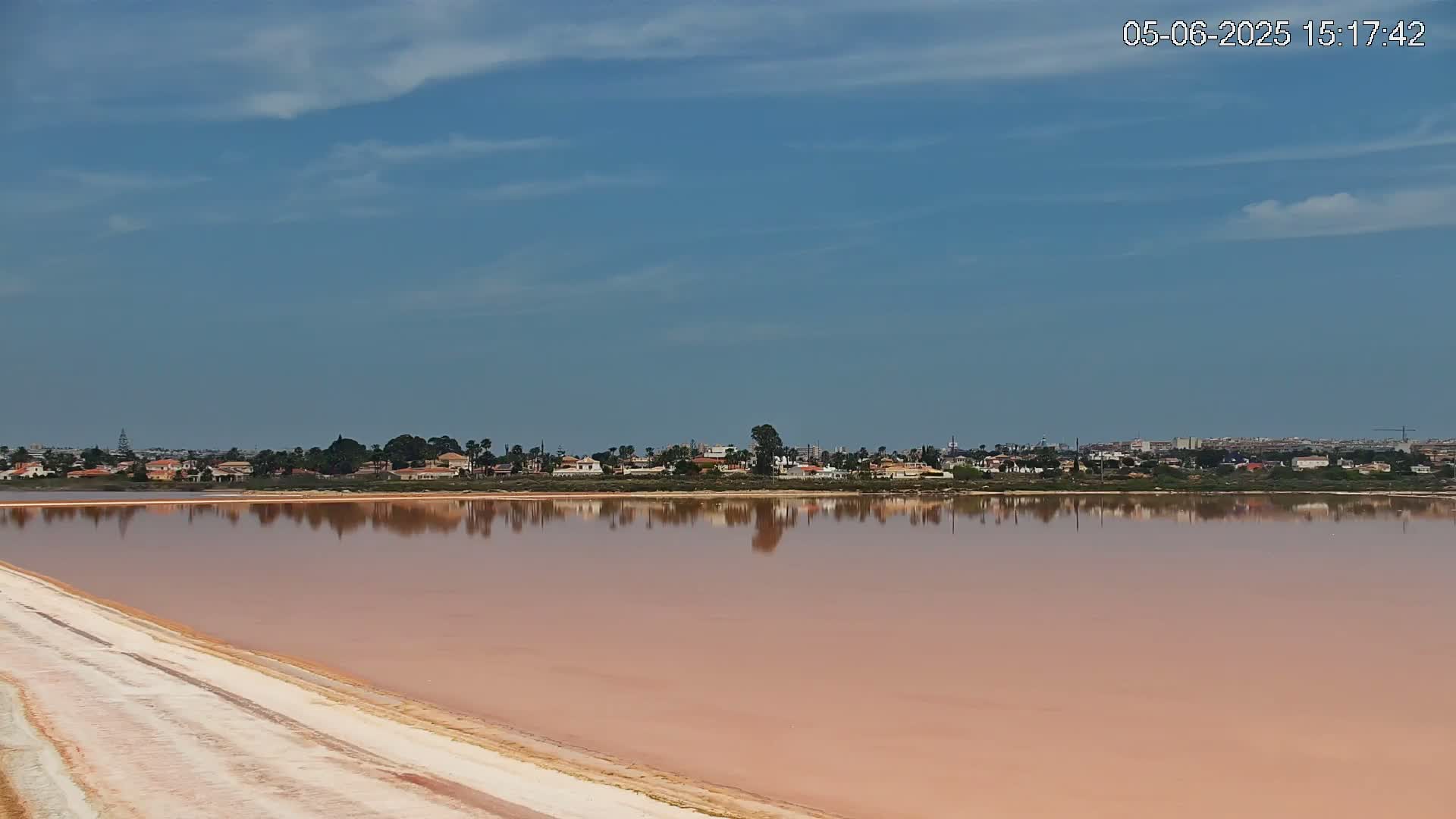 A pink salt lake reflects a distant residential area under a mostly clear, blue sky.