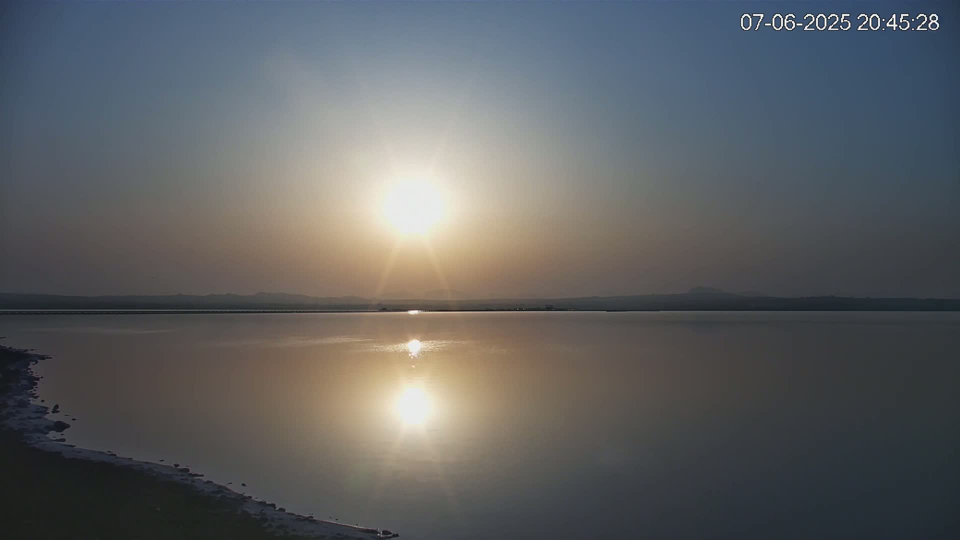 The sun sets over a calm lake, reflecting brightly on the still water, with a distant, hazy shoreline.