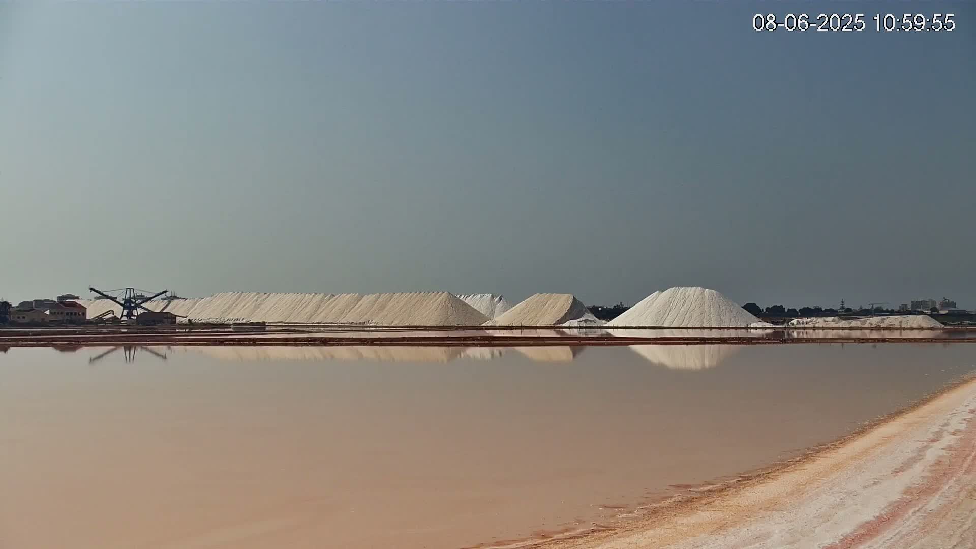 Large piles of salt are reflected in a calm, pinkish-brown body of water under a clear, light-blue sky.