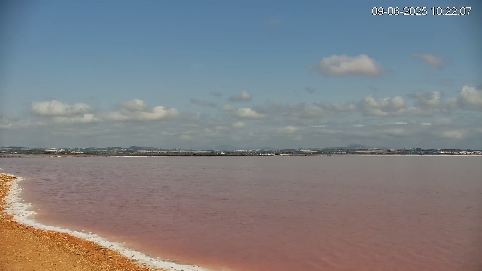 A pinkish-red lake stretches to a distant shoreline under a partly cloudy sky.