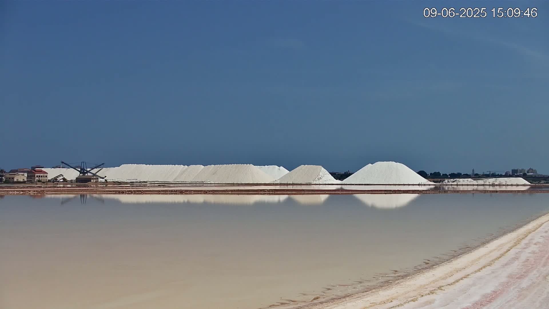 Under a clear blue sky, several large piles of white salt are reflected in a calm body of water next to salt harvesting machinery.