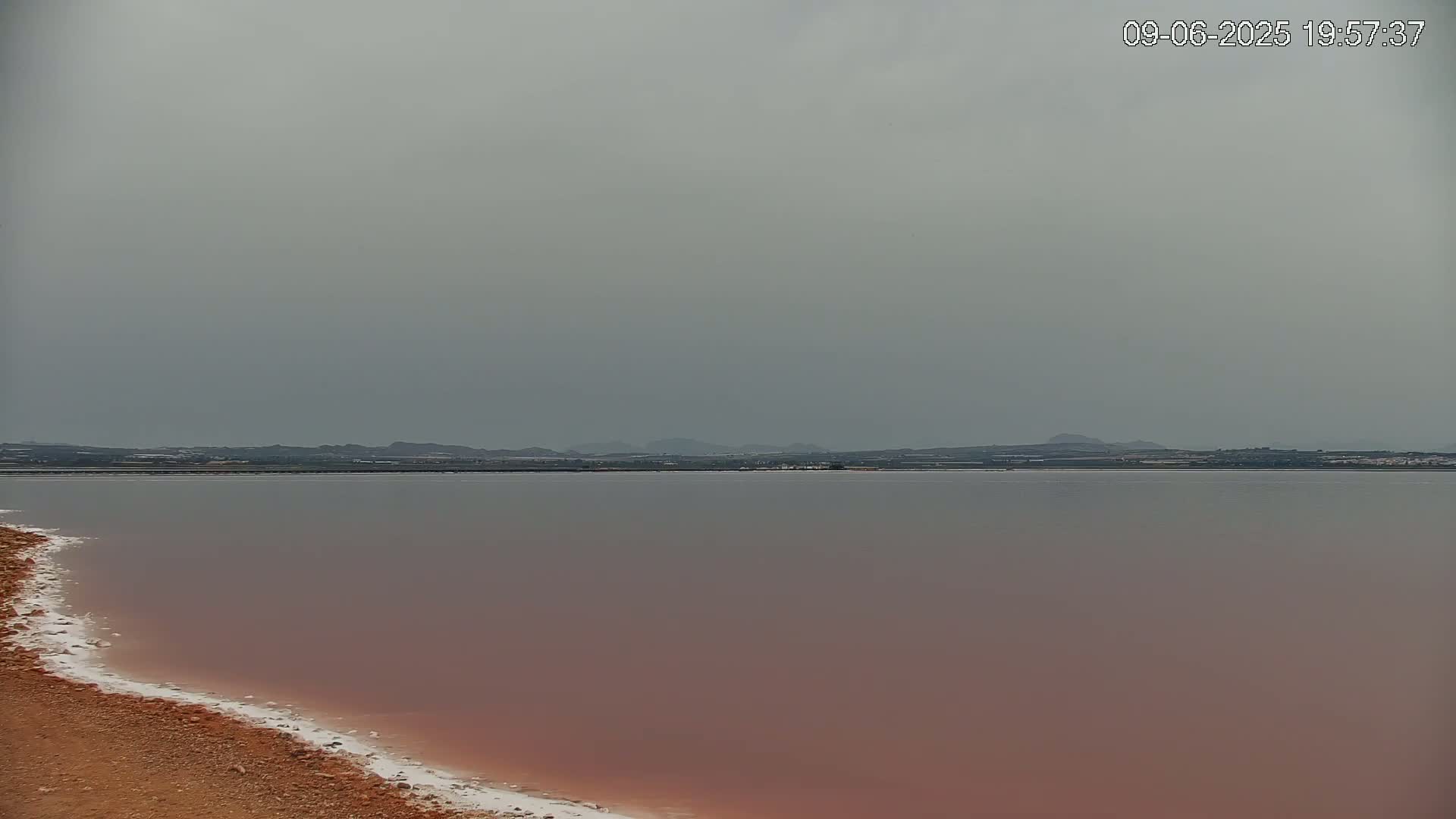 A pinkish-red lake with a white, salty shoreline stretches to a distant, flat land under an overcast sky.