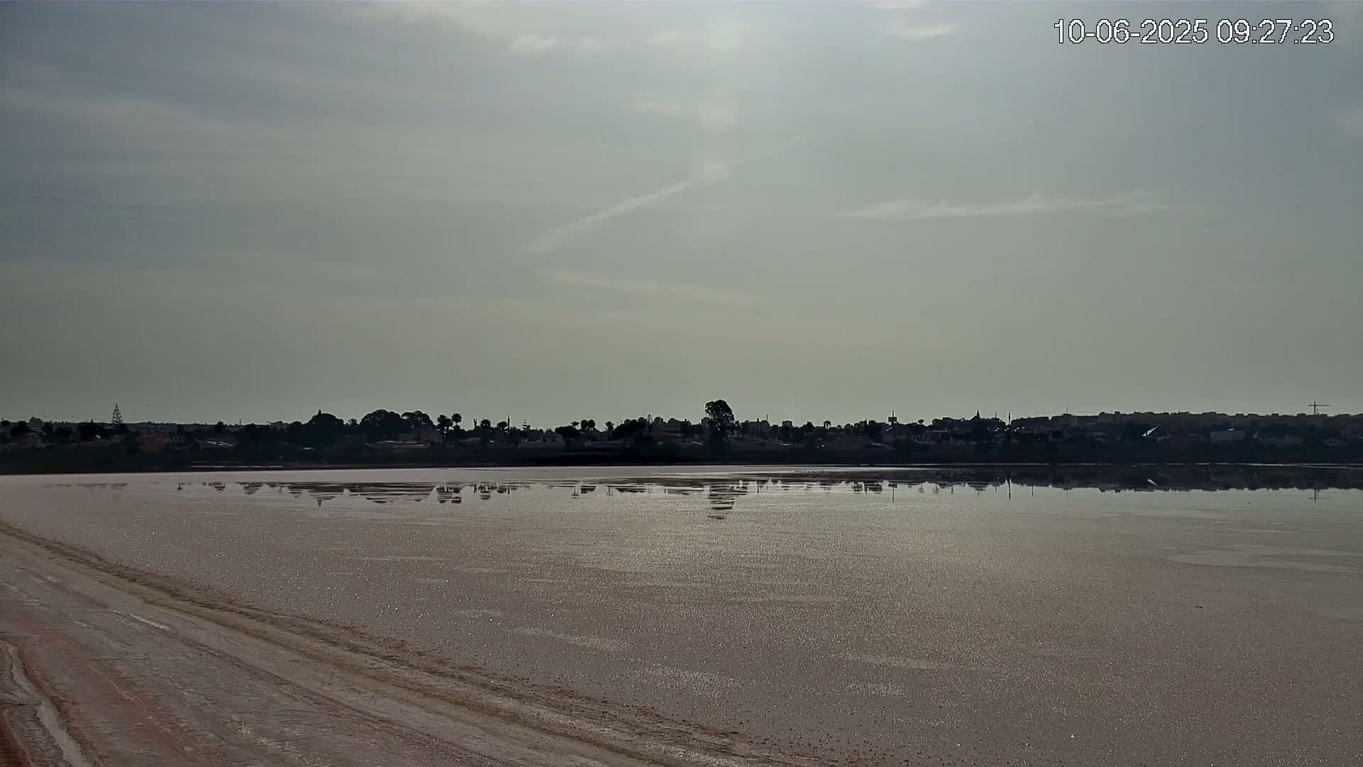 A calm, light-grey-skied day reveals a flat, light-pink lake reflecting a distant, silhouetted town.