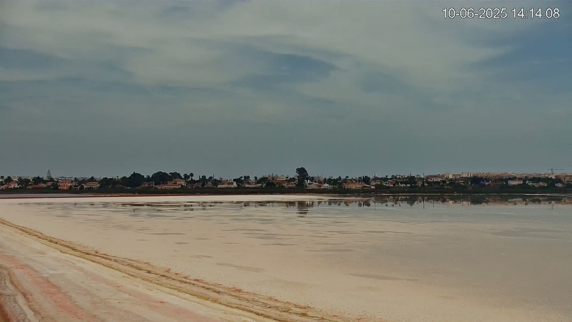 A pale, still salt lake reflects a distant town under a mostly cloudy sky.