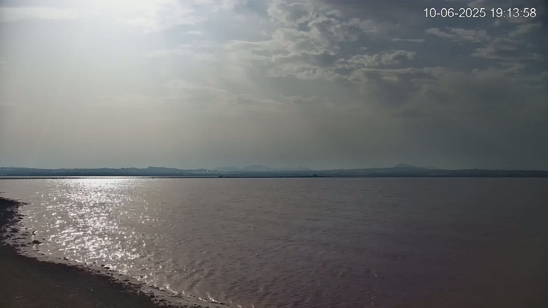 A pinkish-hued body of water reflects sunlight under a partly cloudy sky, with a distant shoreline visible.