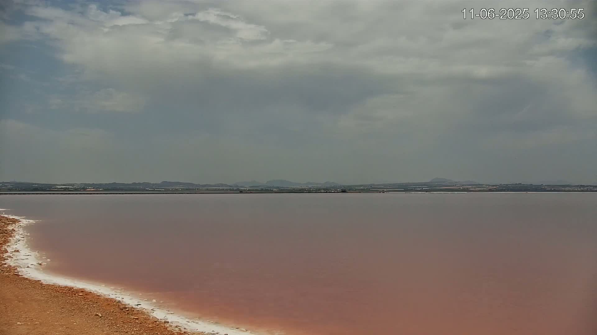 A pinkish lake stretches to a distant, hazy horizon under a mostly cloudy sky.