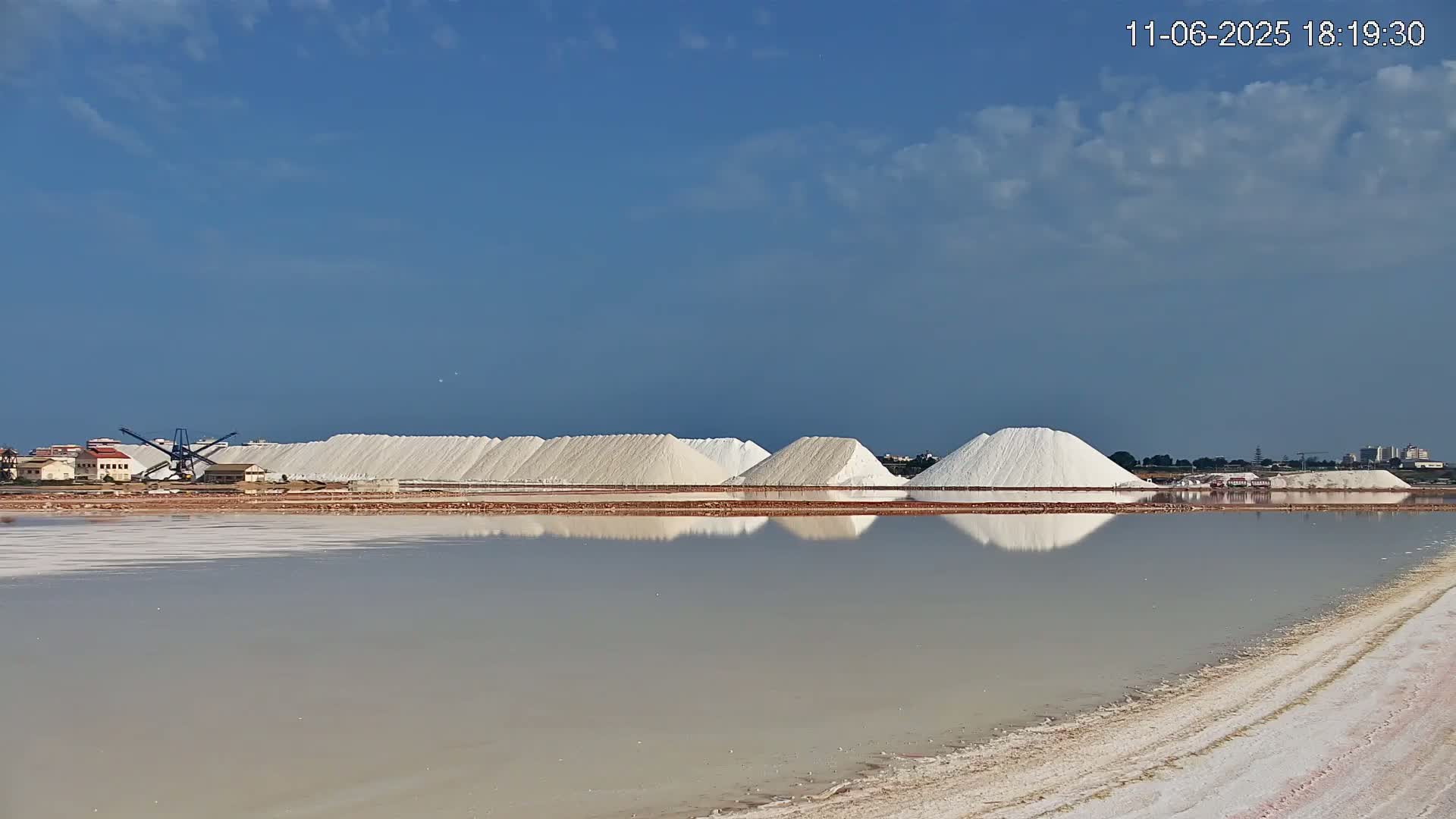 Large piles of white salt are reflected in a calm body of water under a mostly clear blue sky.