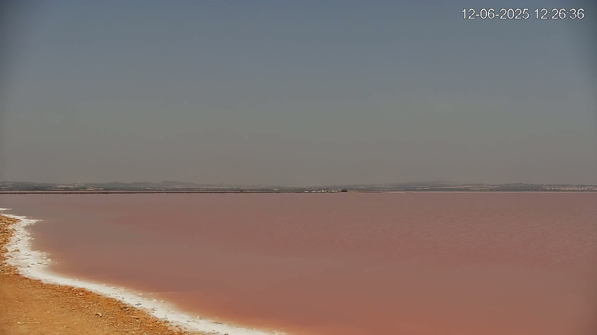 A hazy sky overlooks a pinkish-red lake with a white, salty shoreline.