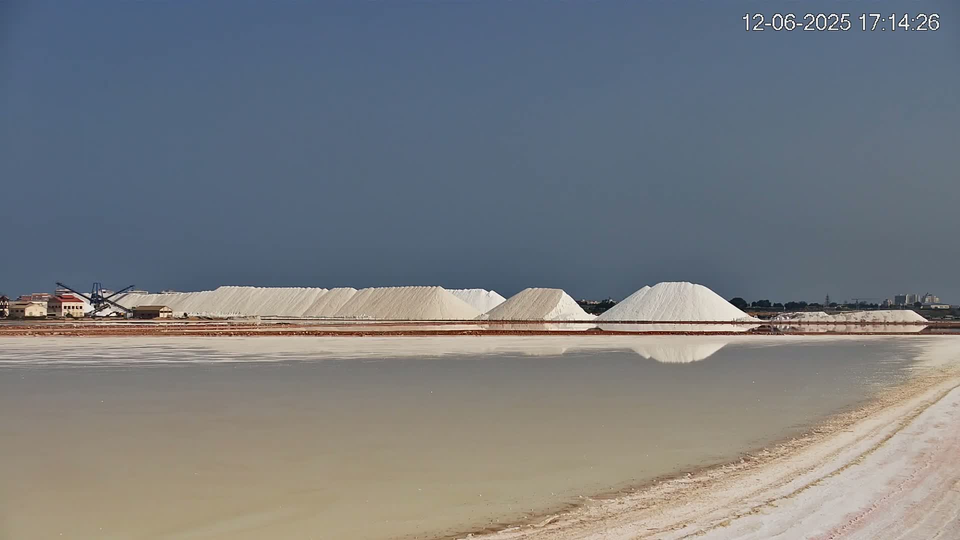 Large piles of white salt sit near a calm body of water under a clear, pale blue sky.