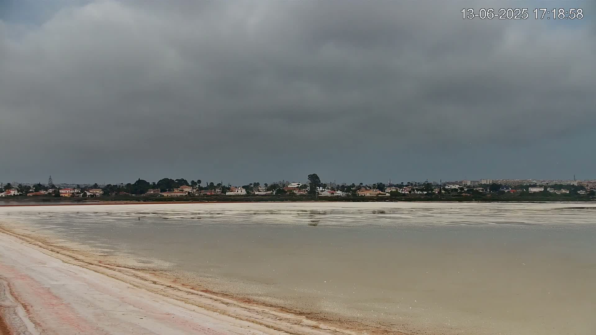 A calm, shallow body of water with a white, salt-crusted surface stretches out to a distant line of houses under a cloudy, overcast sky.
