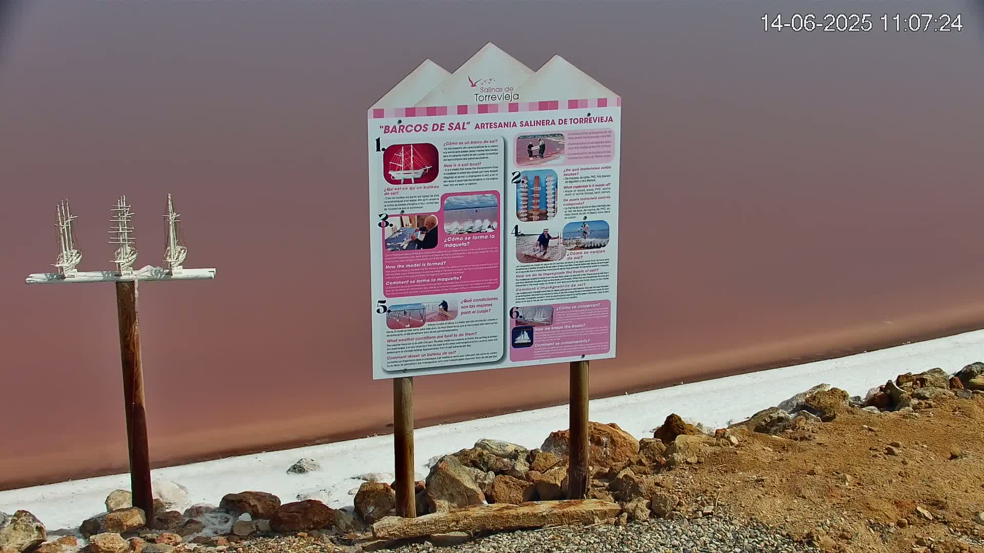 A pink salt lake is visible in the background behind a sign with informational graphics and three small model sailing ships mounted on a post.