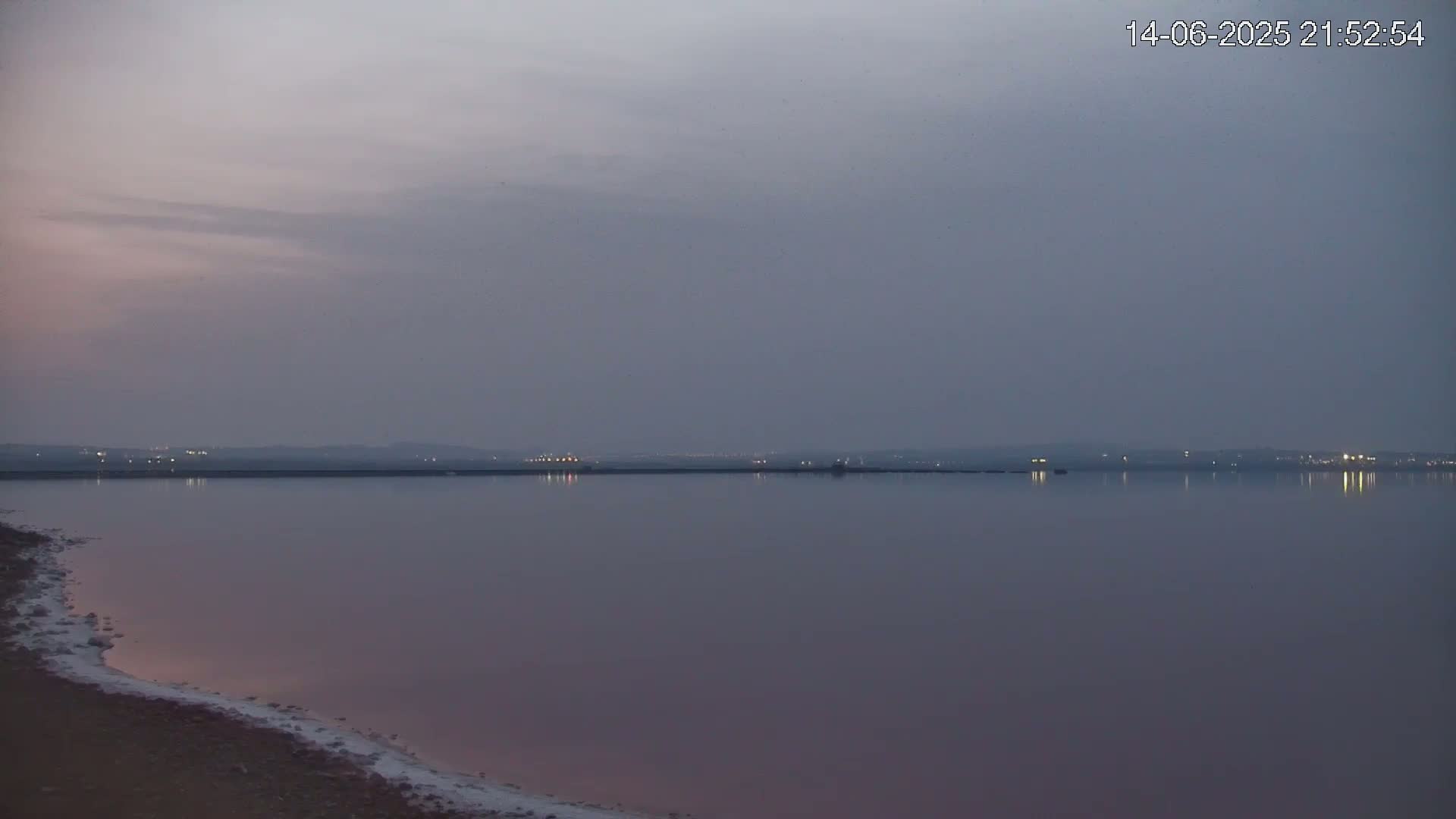 A pink body of water stretches to a distant, hazy shoreline under a cloudy sky.