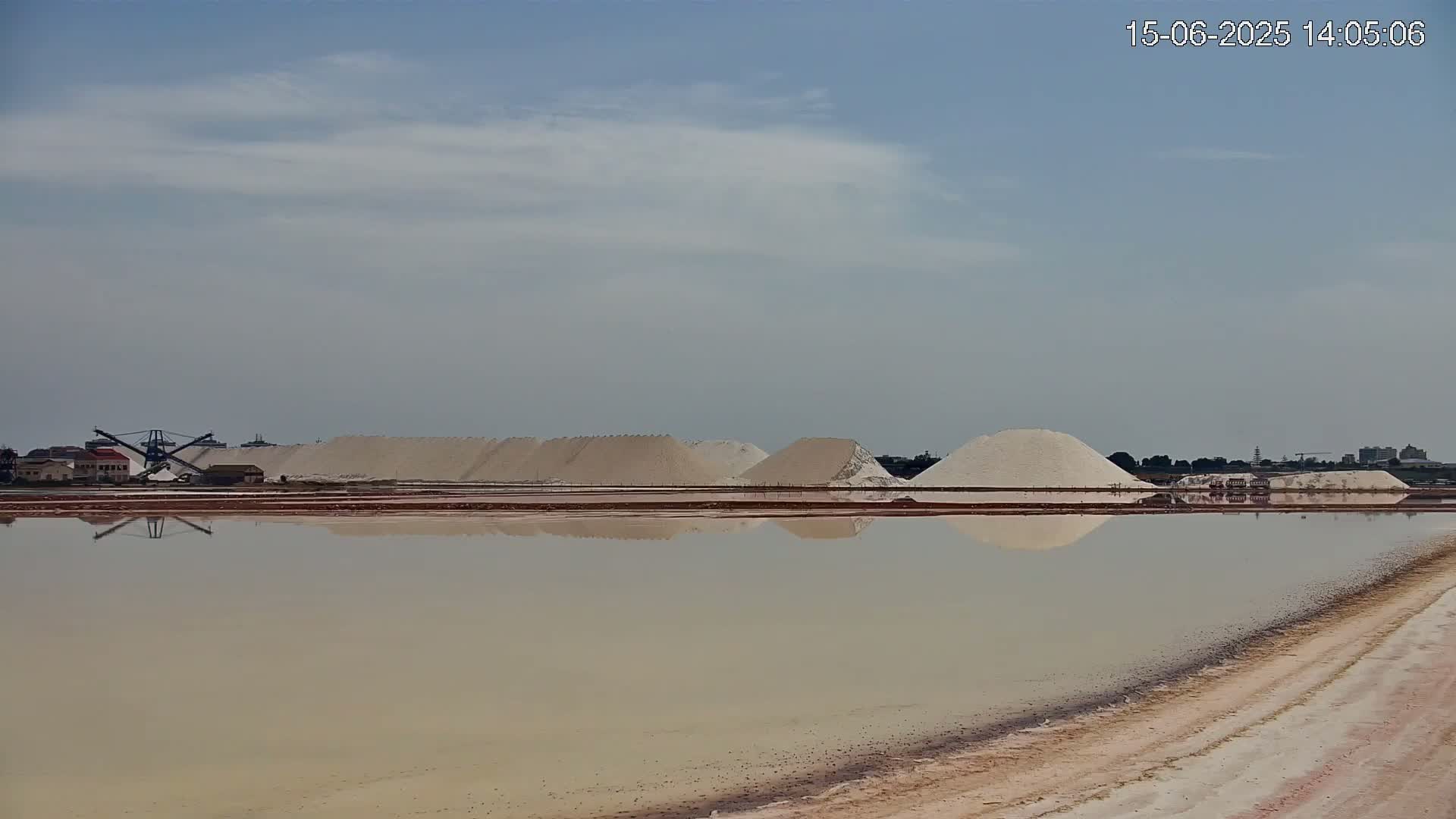 Large piles of white salt are reflected in a calm body of water under a mostly clear sky.