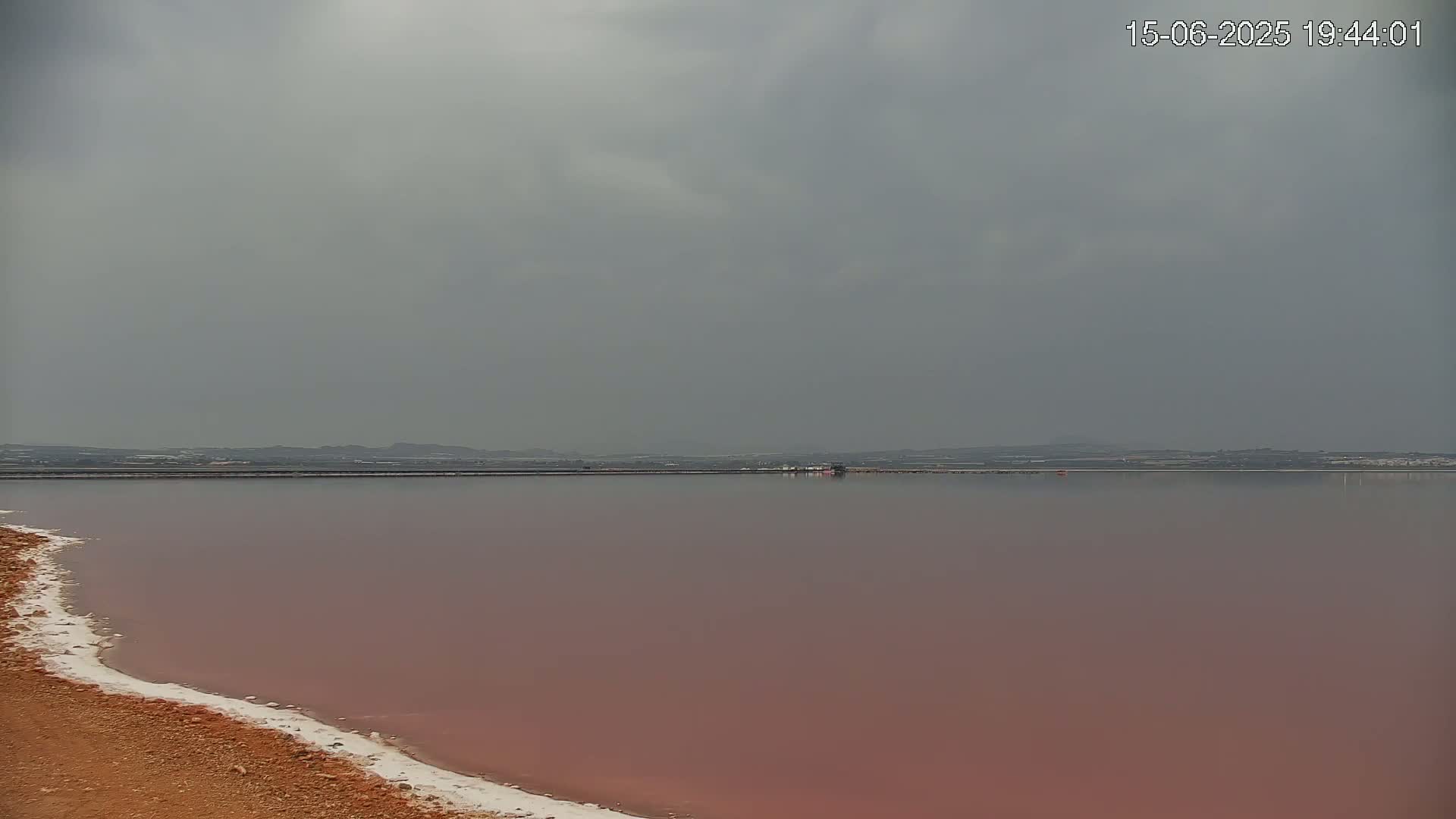A pinkish lake with a white salt edge stretches to a distant shoreline under an overcast sky.