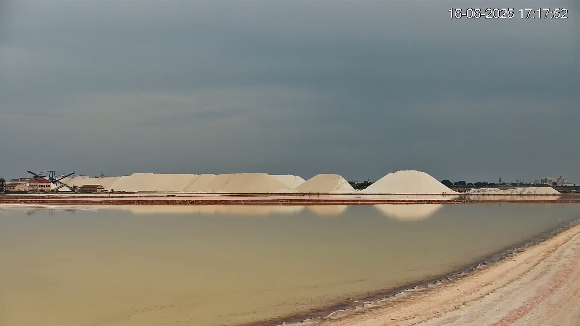 Under a cloudy sky, a calm body of water reflects numerous piles of white salt situated near industrial structures and a shoreline of pale sand.