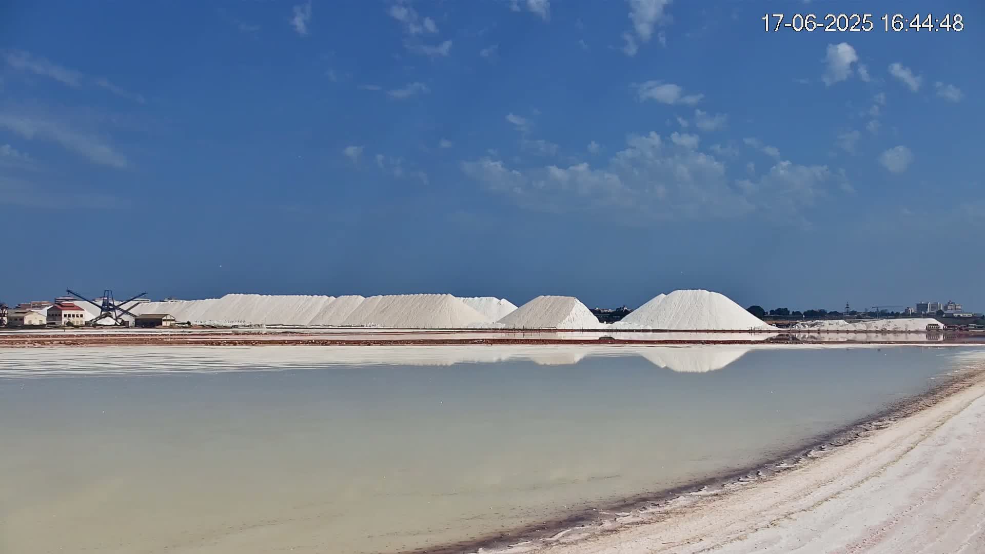Large piles of white salt are reflected in a calm body of water under a mostly clear, blue sky with a few clouds.