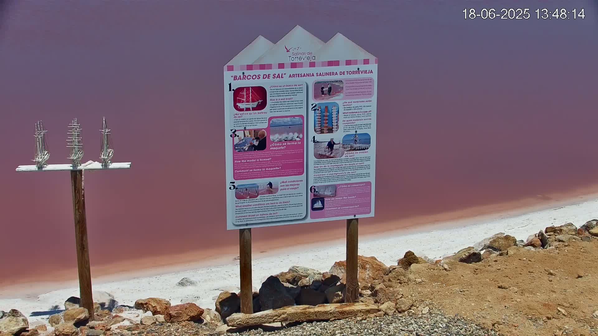 A pink salt lake is bordered by a rocky shore with a wooden sign displaying information about salt boats, and three small model salt boats on a post in the foreground.