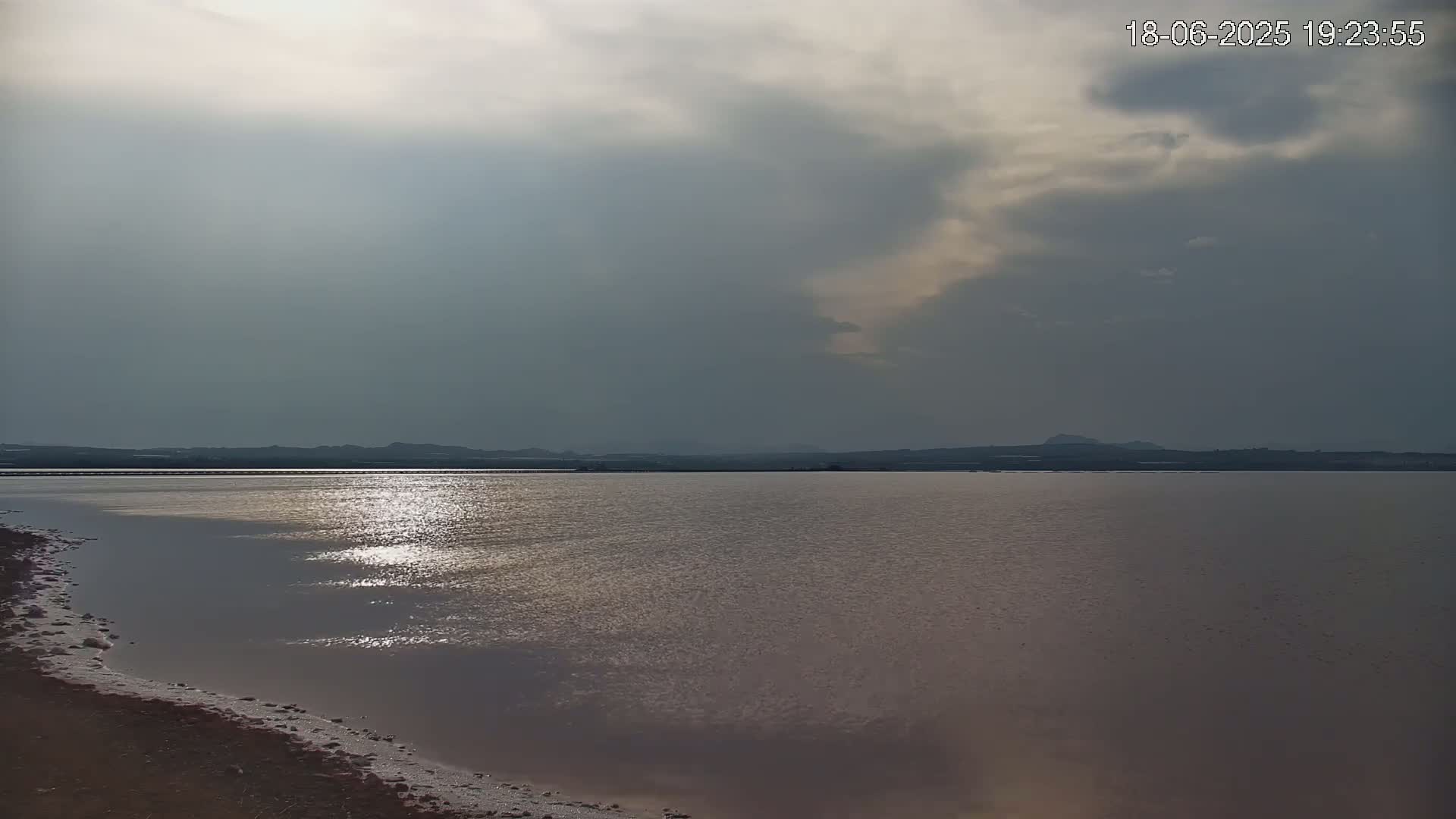 A calm, pink-tinged body of water reflects the sunlight under a mostly cloudy sky.