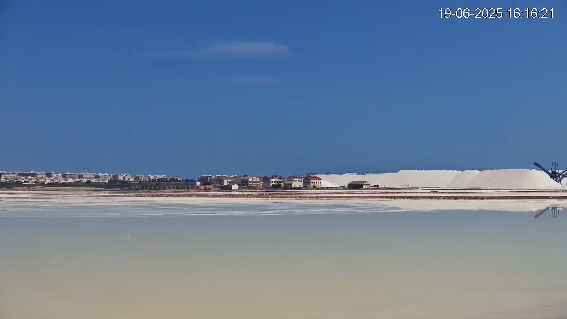 Under a clear blue sky, a large salt flat reflects a distant industrial area with buildings and piles of harvested salt.