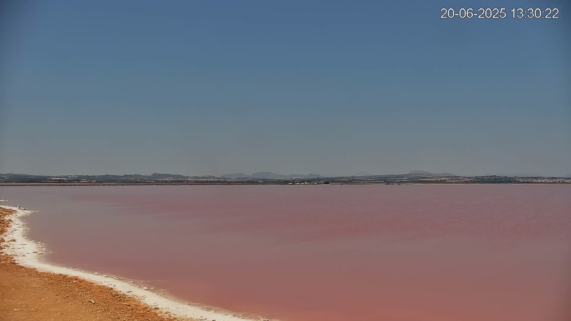 A pinkish-red lake stretches to a distant shoreline under a clear blue sky.