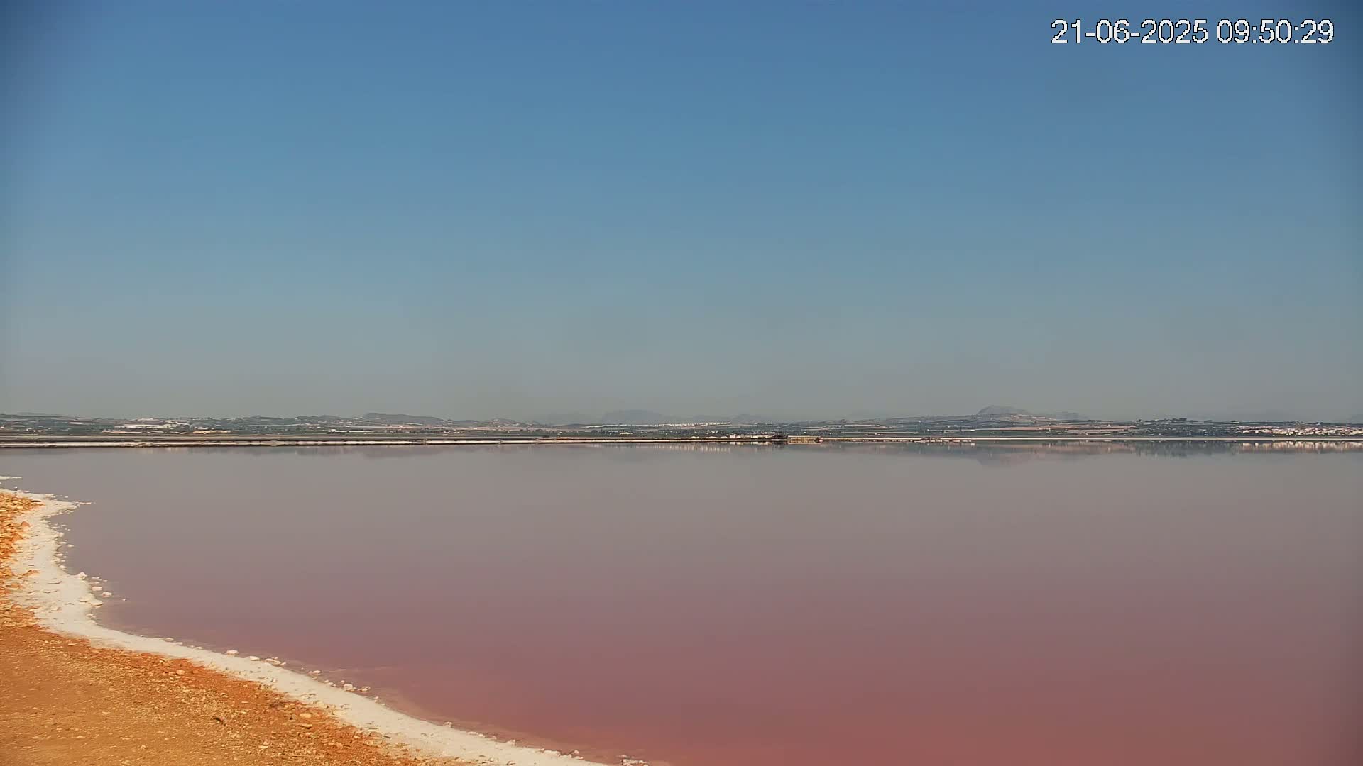 A pinkish lake with a white, sandy shore stretches to a distant horizon under a clear blue sky.