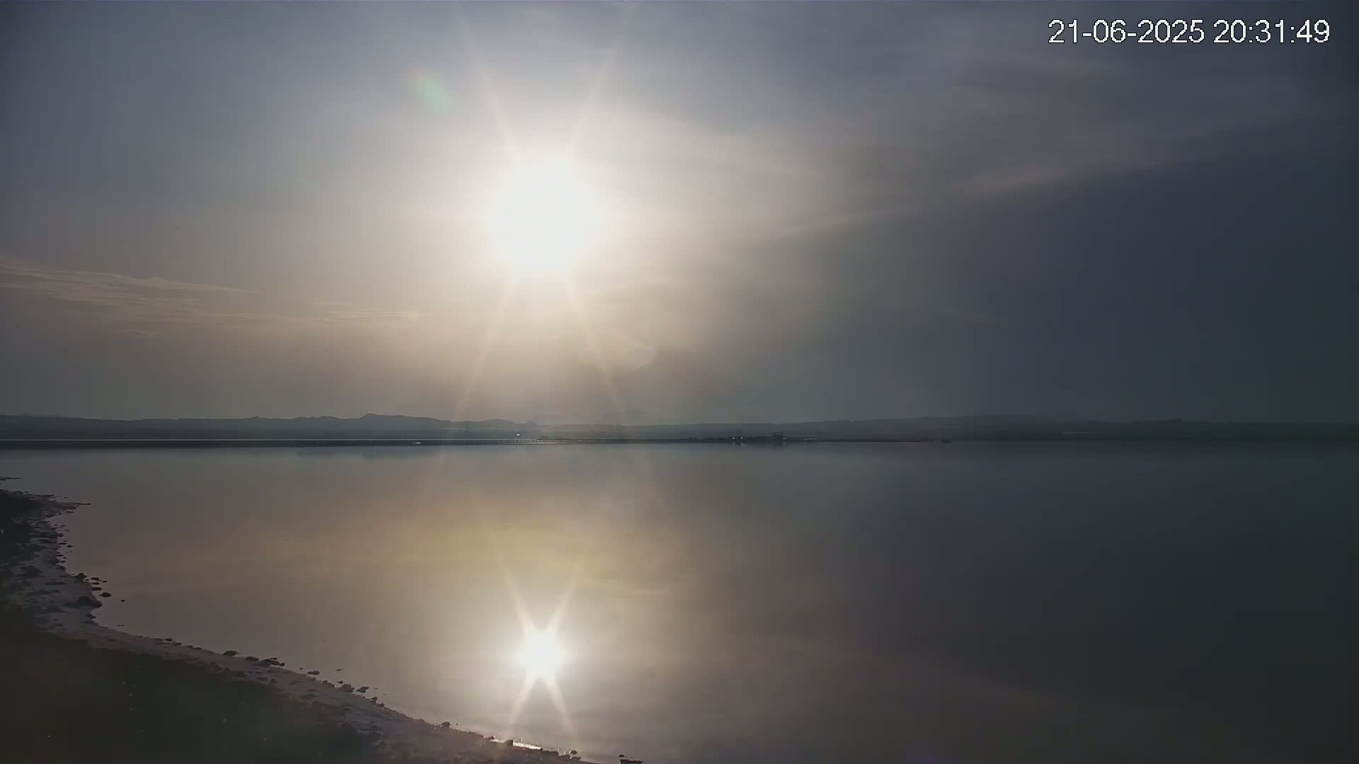 A calm lake reflects a bright sun in an overcast sky, with a distant shoreline visible.