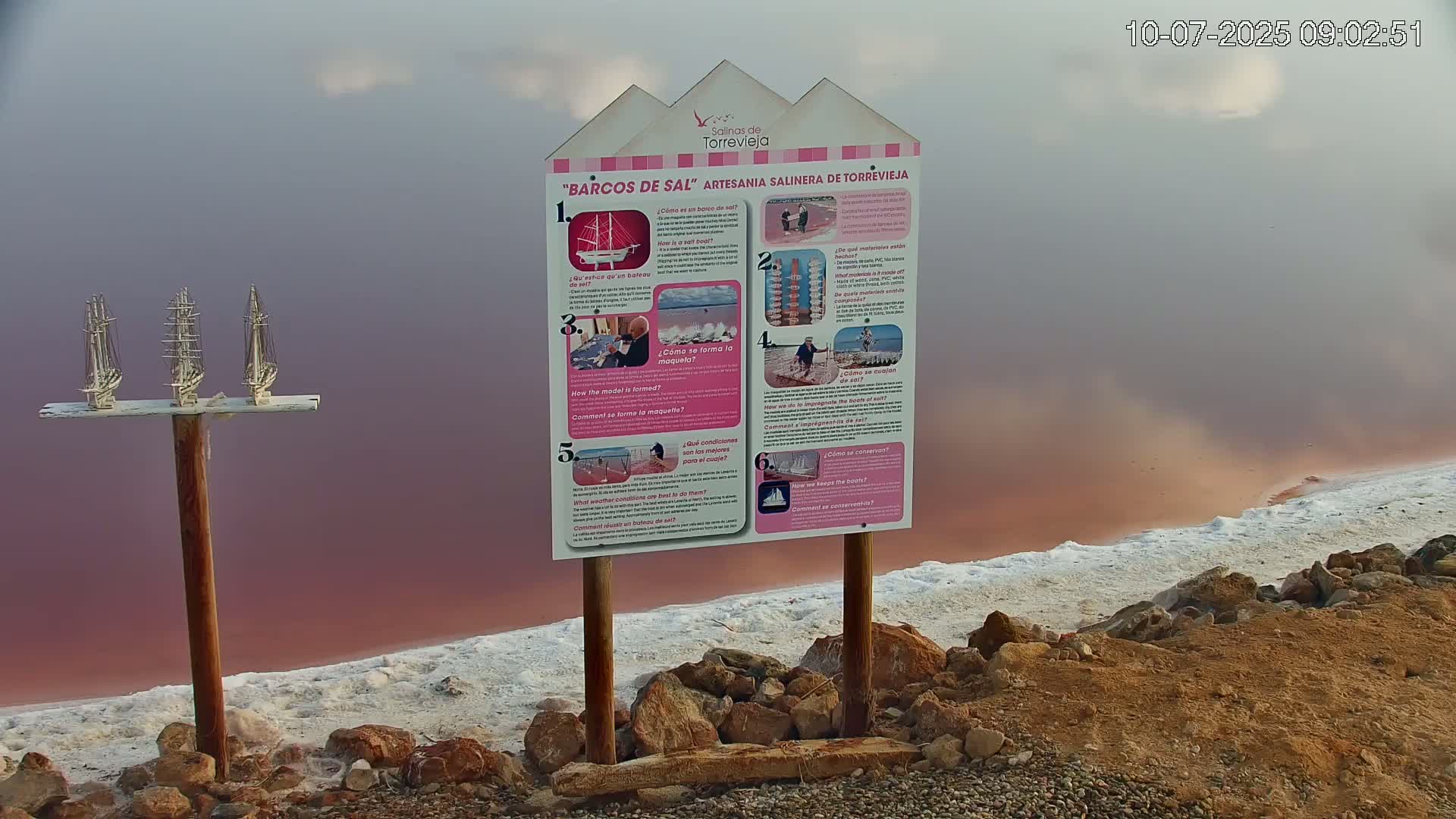 Three miniature sailboats sit atop a post near a pink salt lake edged with white salt deposits under a hazy sky.